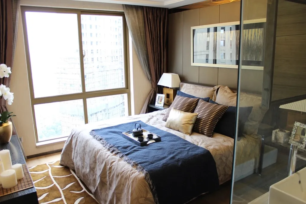 Quarto arrumado, com a cama feita e as almofadas alinhadas, iluminado pela janela